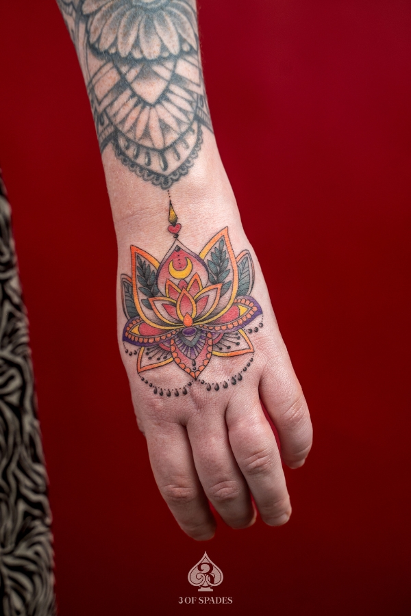 Colorful lotus mandala tattoo on the back of the hand with orange, red, purple, and green detailing – done at 3 Of Spades Tattoo Studio in Hanoi.
