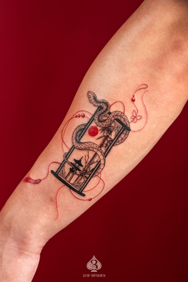 Forearm tattoo of a snake wrapping around an hourglass with Asian scenery and red thread details – done at 3 Of Spades Tattoo Studio, Hanoi.