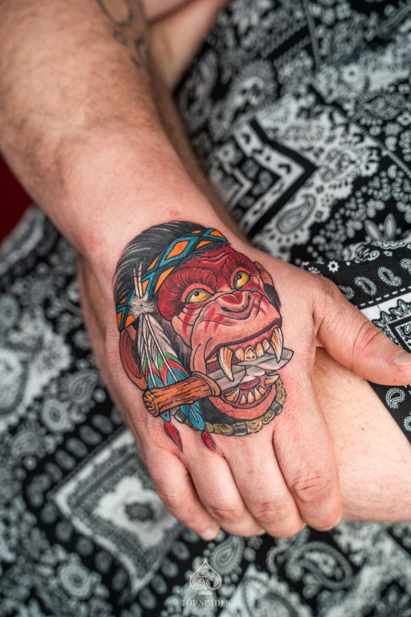 Color tattoo of a fierce gorilla wearing a tribal headdress and biting a bone, inked on the back of the hand at 3 Of Spades Tattoo Studio in Hanoi.