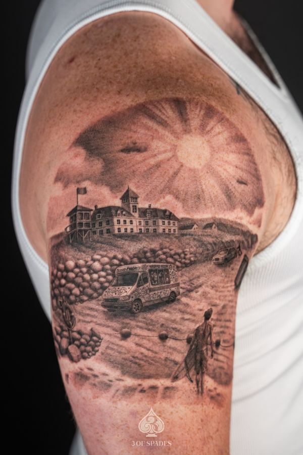 Realistic black-and-grey tattoo of a beach scene with ice cream truck, surfer, and hotel under the sun on upper arm.