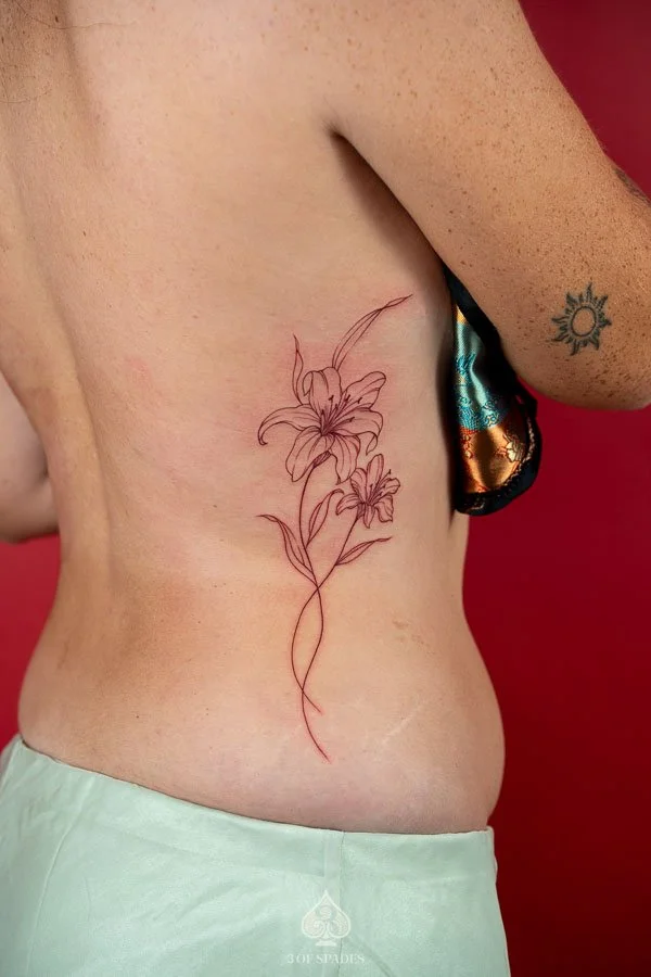 Fine Line Red Lily Flower Hip Tattoo featuring two delicate red ink lily blossoms, flowing stems, and minimalist botanical linework extending from the side ribs toward the hip at 3 of Spades Hano