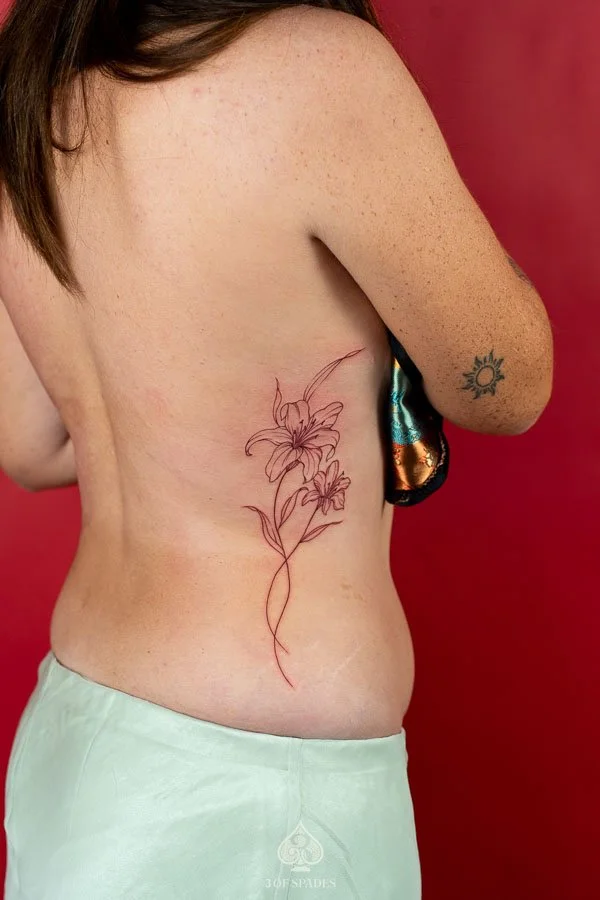 Fine Line Red Lily Flower Hip Tattoo featuring two delicate red ink lily blossoms, flowing stems, and minimalist botanical linework extending from the side ribs toward the hip at 3 of Spades Hano