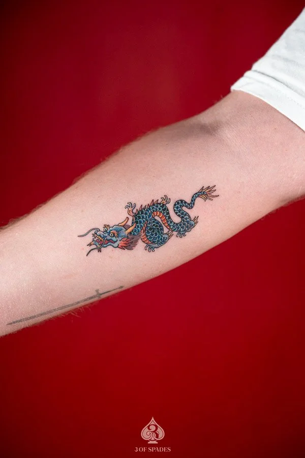 A colorful small old school dragon tattoo featuring a coiled blue dragon, vibrant orange accents, and bold traditional linework.